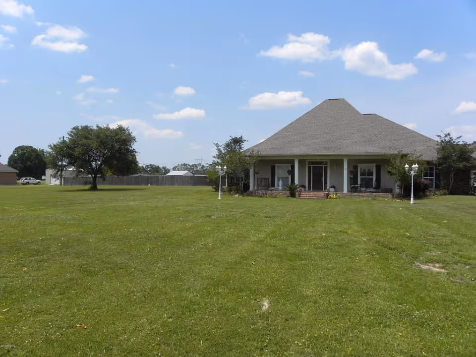 Single-story house with a wide front porch set behind a large grassy lawn under a blue sky.