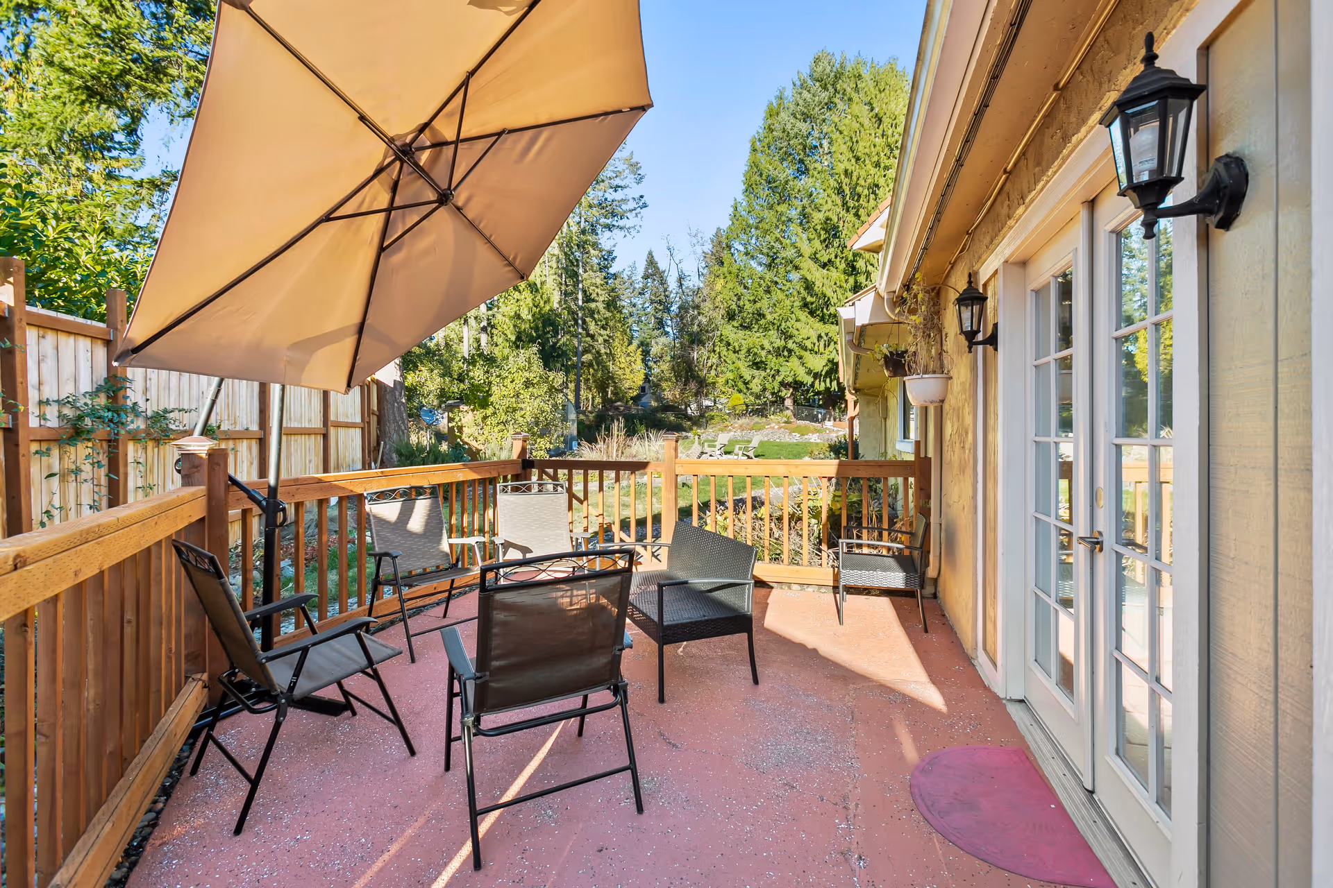 Outdoor patio area with several black metal chairs arranged around a large beige umbrella. The patio has a reddish floor and wooden railing. There are two black wall-mounted lanterns on the beige exterior wall of the building with white-framed glass doors leading inside. In the background, there are green trees and a grassy area with additional seating.