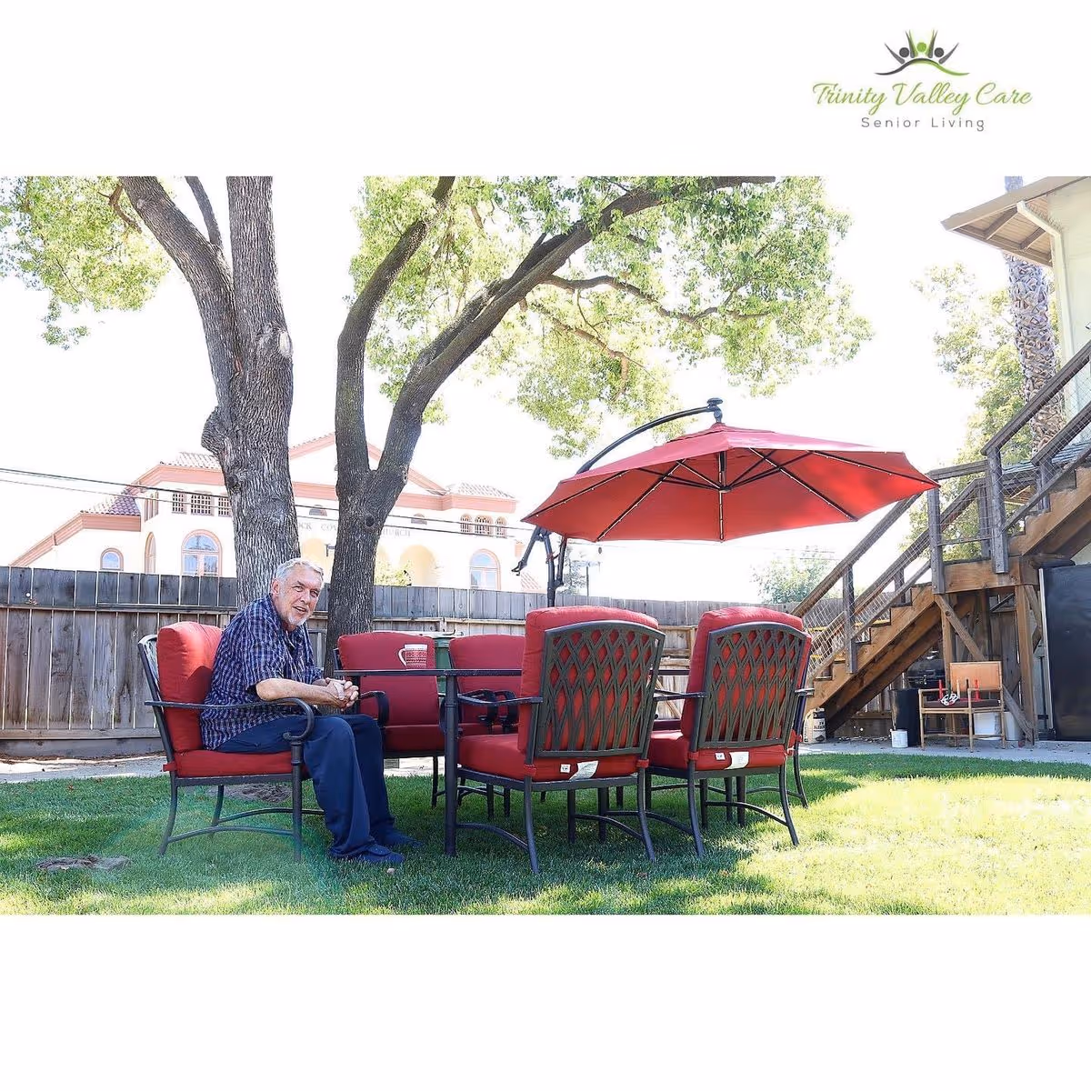 An older man sits at a red patio dining set under a red umbrella in a grassy backyard with trees and wooden stairs.