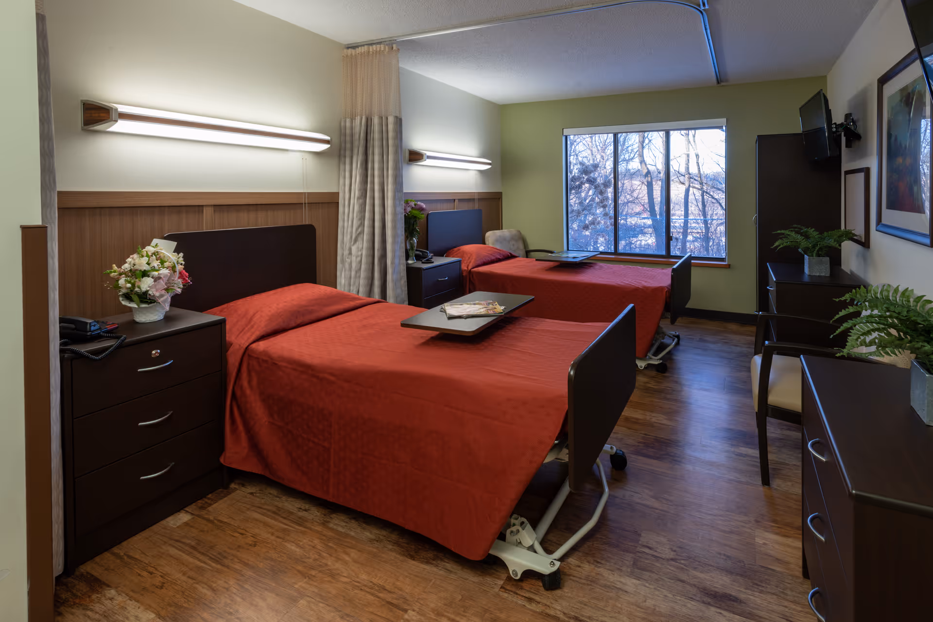 A well-lit room with two hospital-style beds covered in red bedspreads, each with an overbed table. The room has wooden flooring, a large window showing trees outside, bedside tables with flowers, wall-mounted lights above each bed, a TV mounted on the wall, and several pieces of dark wood furniture including dressers and chairs.