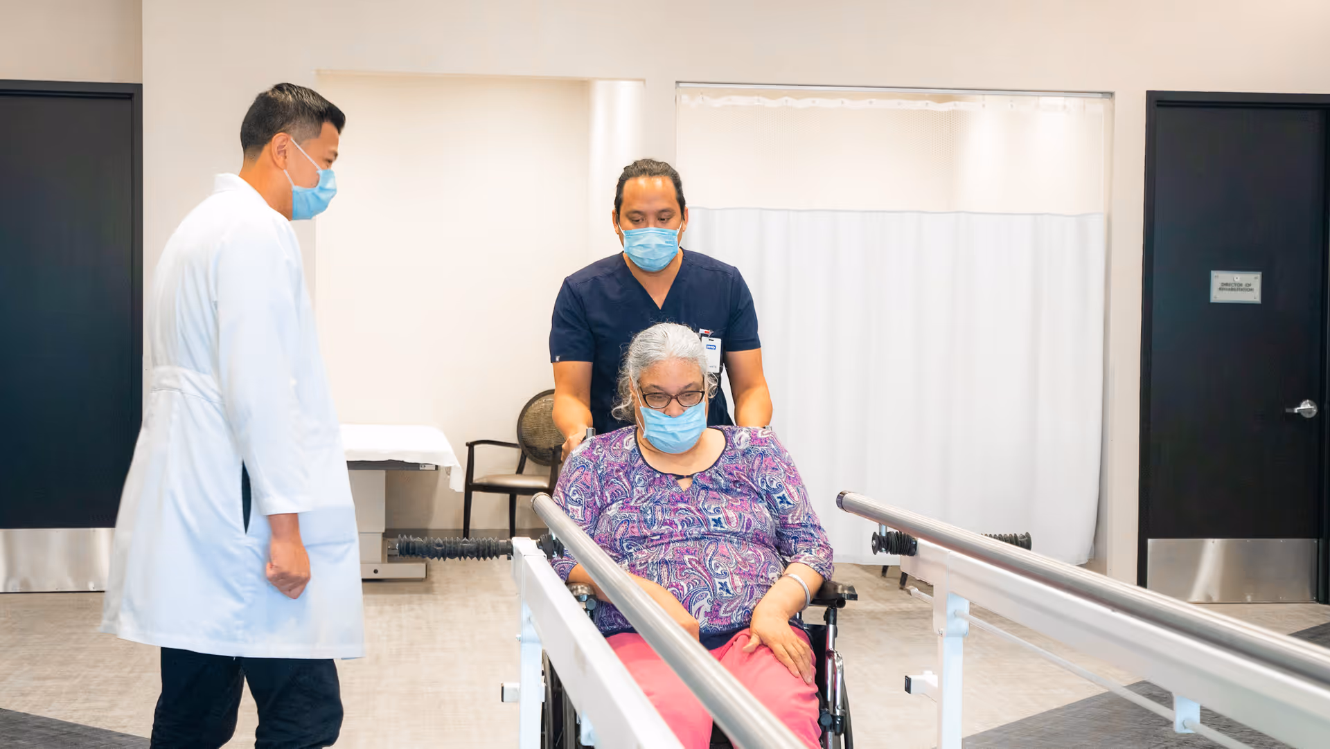 A healthcare worker in navy scrubs pushes an elderly woman in a wheelchair along parallel bars in a rehabilitation area. Another healthcare professional in a white coat observes. All three individuals are wearing face masks.