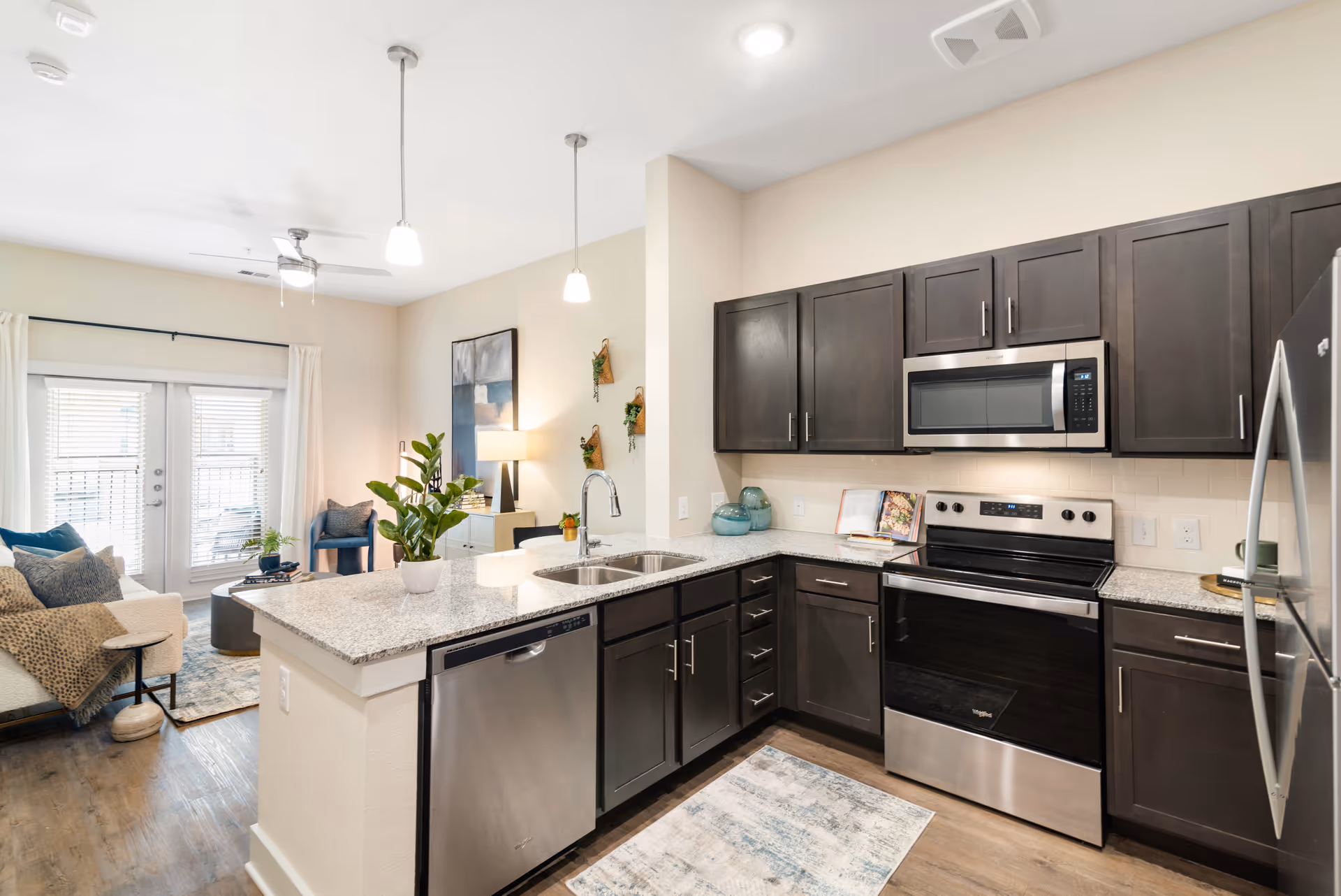 Modern kitchen with dark wood cabinets, stainless steel appliances including a dishwasher, oven, microwave, and refrigerator. Granite countertops with a double sink and a potted plant on the island. The kitchen opens into a living area with a sofa, chair, coffee table, and large windows with white curtains allowing natural light.