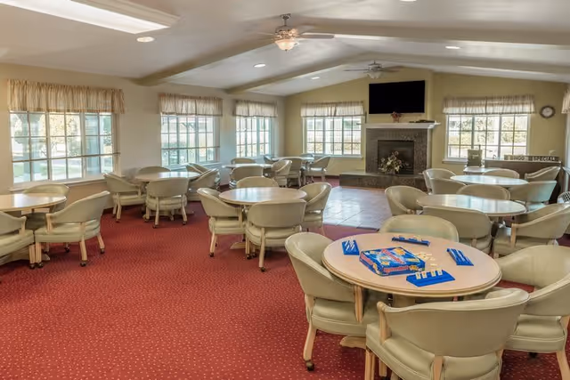 A spacious common room with multiple round tables and beige cushioned chairs arranged around them. The room has large windows with beige valances allowing natural light to fill the space. A fireplace with a mounted flat-screen TV above it is centered on one wall. The floor is covered with red carpet, and a ceiling fan is visible. One table in the foreground has a board game set up on it.