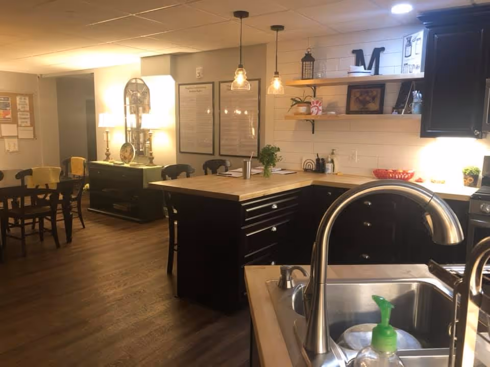 Interior view of a kitchen and dining area in an assisted living facility. The kitchen features a sink with a modern faucet, black cabinets, wooden countertops, and open shelves with decorative items. Pendant lights hang above the kitchen island. In the background, there is a dining table with chairs and a sideboard with lamps and framed pictures on the wall.