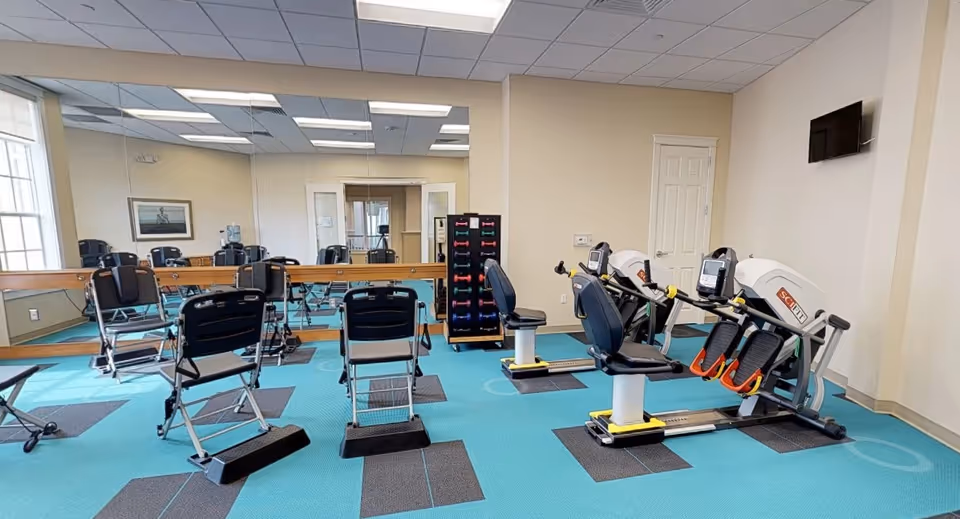 A fitness room with exercise equipment including two seated rowing machines, several black folding chairs, a rack of colorful dumbbells, a large mirror on the wall, a wall-mounted TV, and a window letting in natural light.