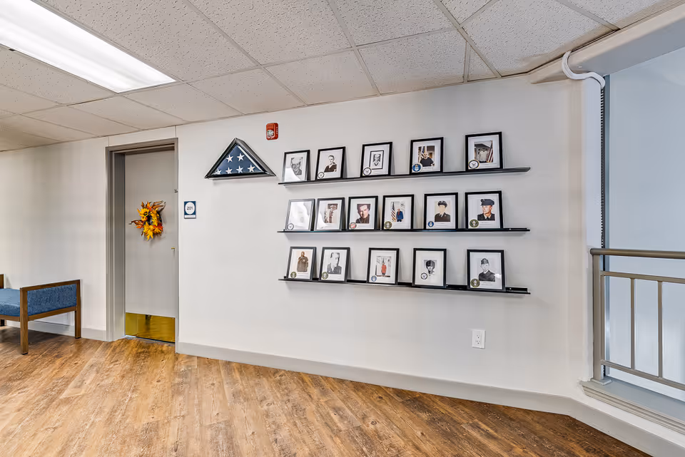 Interior hallway with framed photos on wall-mounted shelves, a folded American flag display, a doorway with a wreath, and a bench.