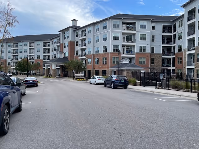 Front view of a multi-story senior living building with balconies, parked cars, and an entrance driveway.