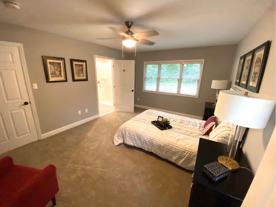 Spacious bedroom with a bed, nightstands and lamps, a ceiling fan, window, and an open door to an en-suite bathroom.