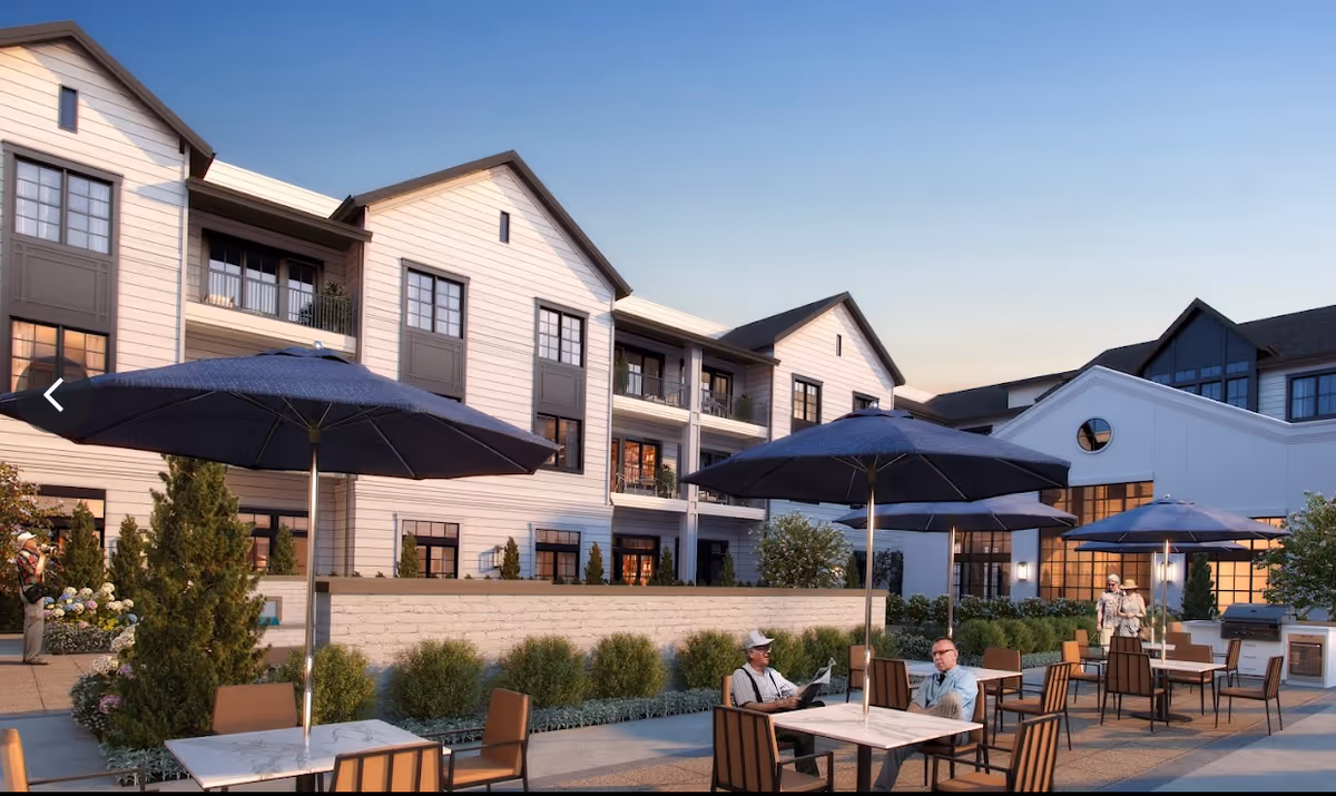 Outdoor patio area at Opus East Memphis with tables and chairs under large blue umbrellas. Two men are seated at one table, and a couple is walking nearby. The background shows a multi-story building with balconies and large windows, surrounded by greenery and bushes.