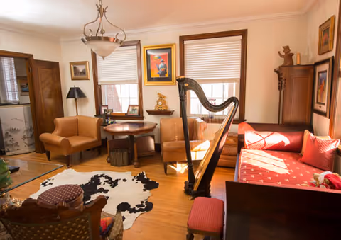 A cozy living room with wooden floors and warm lighting featuring a red cushioned bench, a harp, two armchairs, a round wooden table, a cowhide rug, and various framed artworks on the walls.