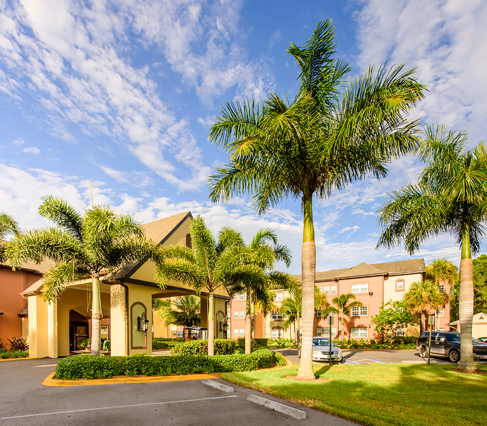 Exterior view of Brookdale College Parkway facility featuring a driveway entrance with a covered portico, surrounded by palm trees and well-maintained landscaping under a partly cloudy blue sky. Several parked cars are visible near the building.