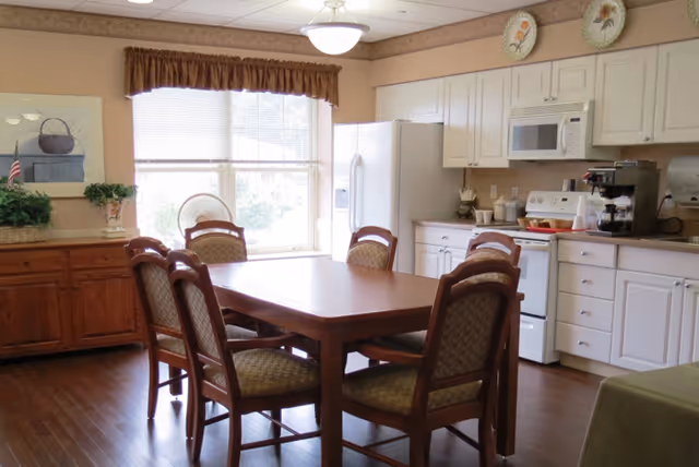 A bright kitchen and dining area with a wooden dining table surrounded by six cushioned chairs. The kitchen features white cabinets, a white refrigerator, a white stove with a microwave above it, and a coffee maker on the counter. There is a window with blinds and a brown valance letting in natural light. The floor is wooden, and there is a wooden sideboard with plants and a framed picture on the wall.