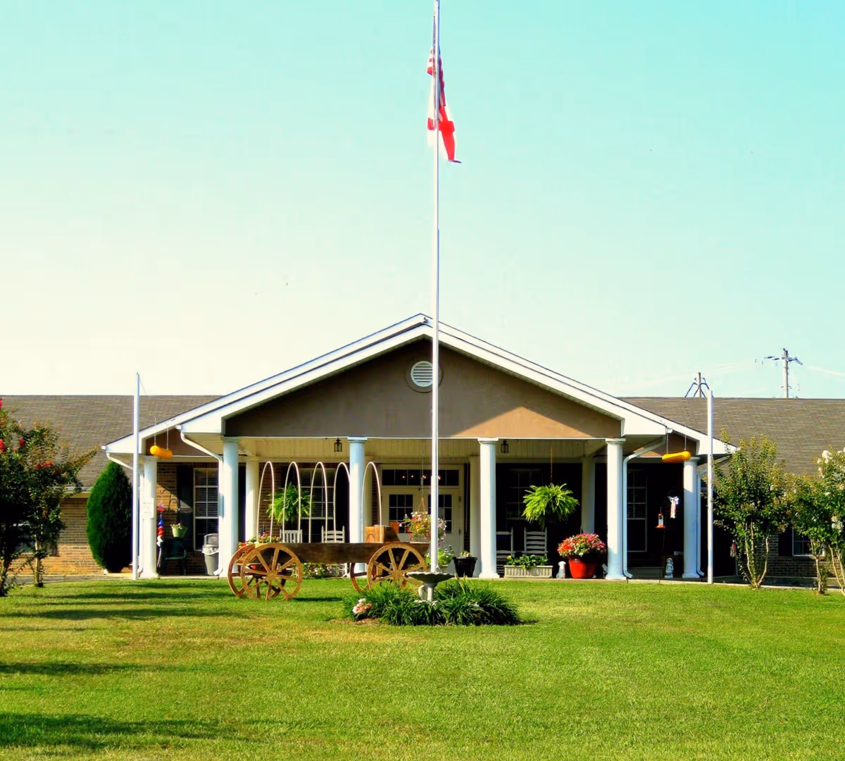 Front exterior view of a single-story building with a covered porch supported by white columns. There is a flagpole with an American flag in front of the building, a wooden wagon decoration on the lawn, and various plants and flowers around the porch area. The sky is clear and blue.