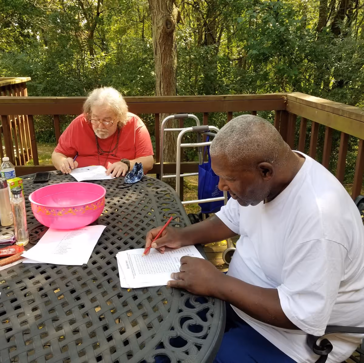 Two elderly men sitting at a metal outdoor table on a wooden deck surrounded by trees. They are engaged in an activity involving papers and pencils, with one man wearing a red shirt and glasses and the other wearing a white shirt. A walker is visible behind them.