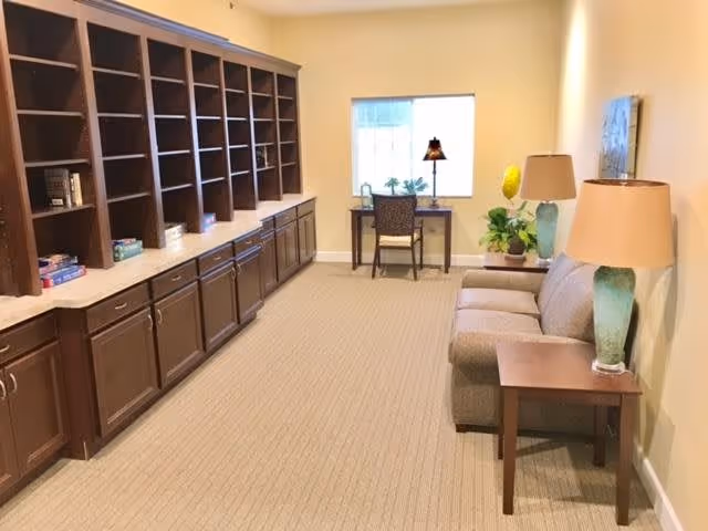 A well-lit interior room with beige walls and carpeted floor. On the left side, there is a long built-in wooden cabinet with multiple open shelves and closed drawers. At the far end of the room, a small desk with a chair is positioned in front of a window, with a lamp and some plants on the desk. On the right side, there is a gray sofa with two side tables, each holding a lamp. There are additional plants and a piece of wall art above the sofa.