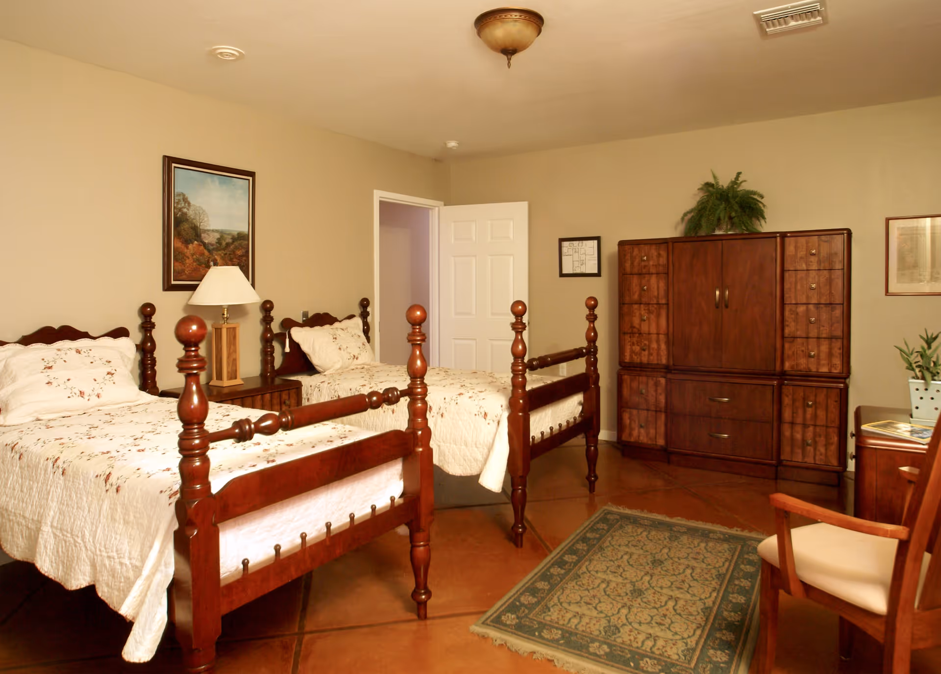 A bedroom with two wooden twin beds with white floral bedding, a wooden nightstand with a lamp between the beds, a wooden dresser with a plant on top, a framed picture on the wall, a patterned rug on the floor, and a wooden chair with a cushion.