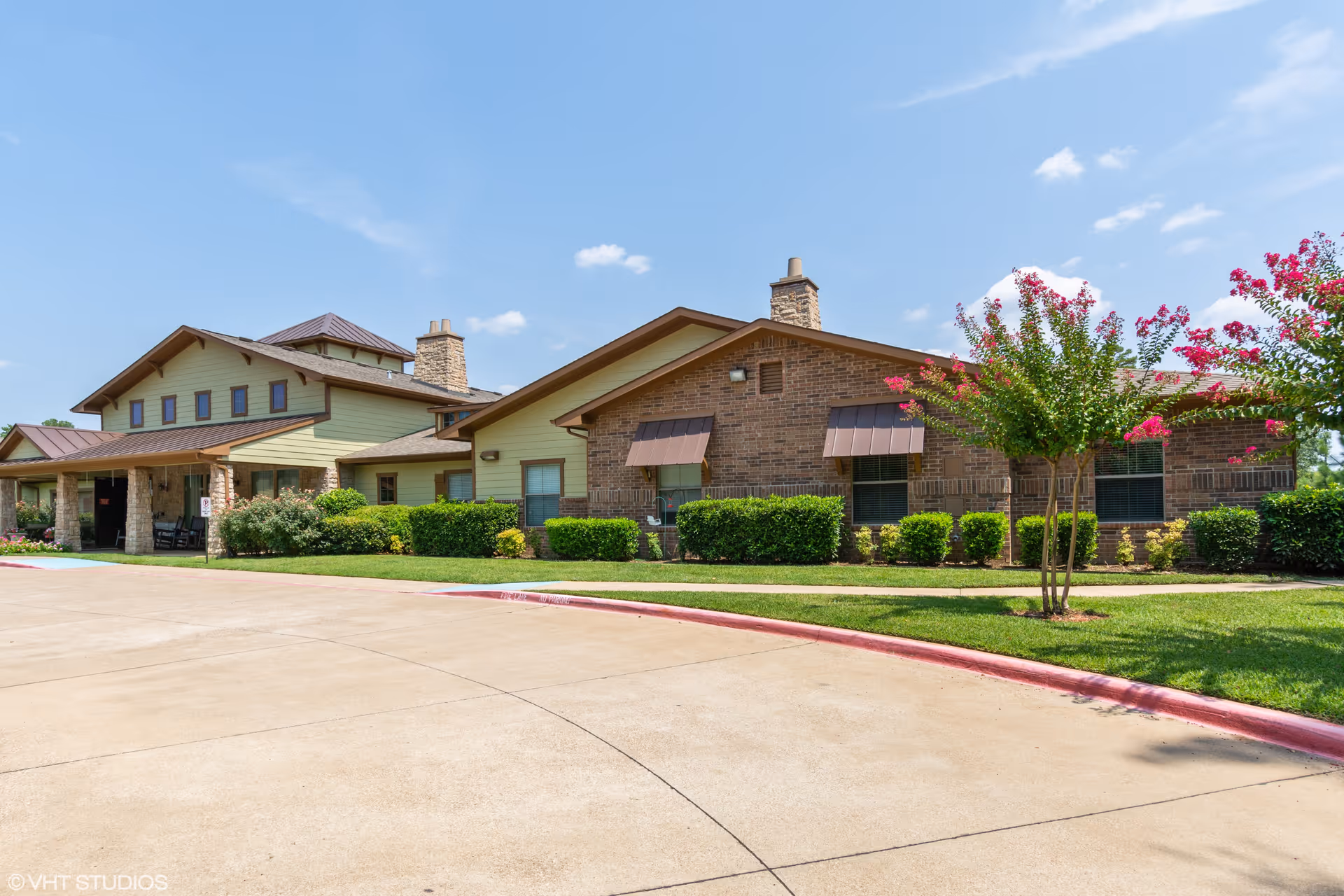 Exterior view of Azalea Trails Assisted Living and Memory Care facility showing a single-story building with brick and light green siding, multiple windows with awnings, a covered entrance with stone pillars, well-maintained landscaping including bushes and flowering trees, and a clear blue sky above.