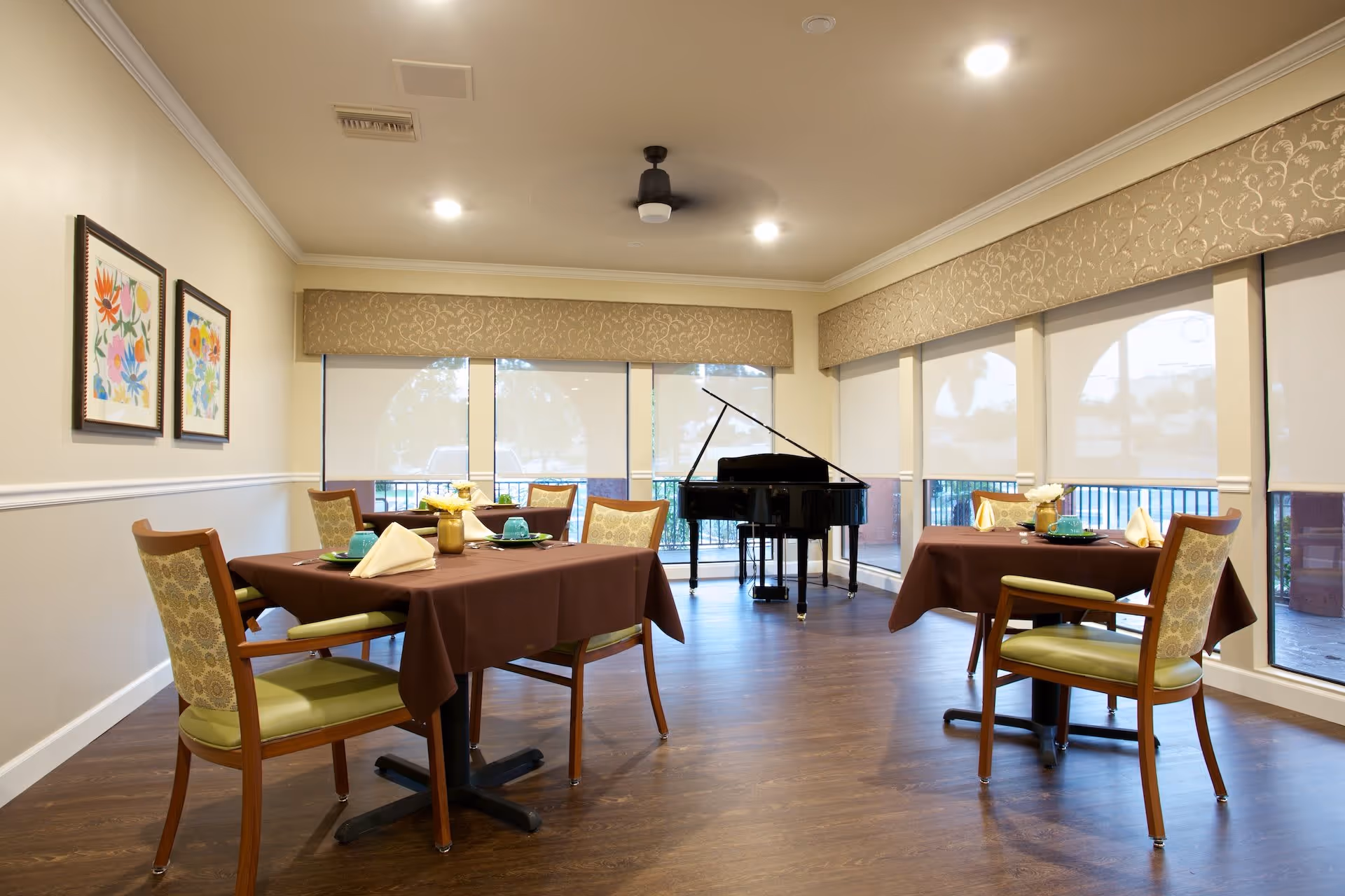 A bright dining room with wooden floors, three tables covered with brown tablecloths, each set with green plates, blue cups, and folded beige napkins. There are wooden chairs with green cushions around the tables. Large windows with beige valances let in natural light. A black grand piano is positioned near the windows.