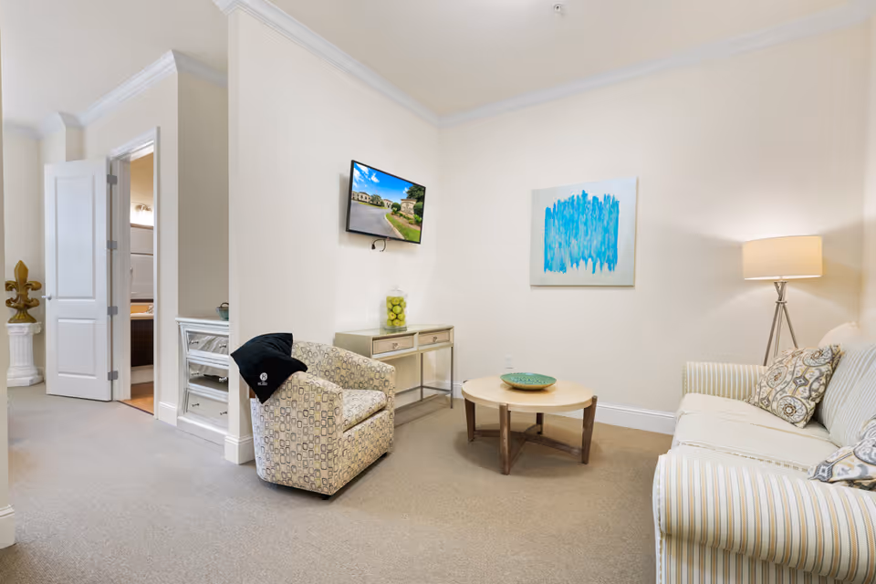 A cozy living room area with a striped sofa, patterned armchair, round wooden coffee table, and a floor lamp. A small console table with a jar of green apples is placed against the wall beneath a mounted flat-screen TV displaying an outdoor scene. A blue abstract painting hangs on the wall. An open door leads to a bathroom.