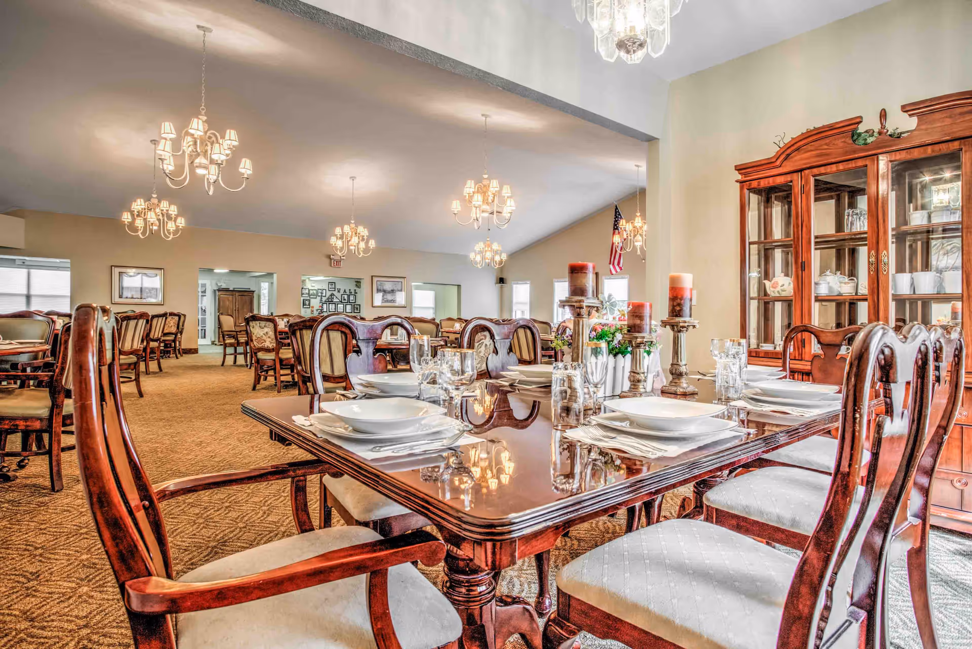 A spacious dining room with multiple wooden tables and chairs arranged neatly. The table in the foreground is set with white plates, bowls, glasses, and silverware, along with decorative candles and flowers. The room is well-lit with several chandeliers hanging from the ceiling, and a wooden china cabinet filled with dishes is visible on the right side. The walls are light-colored, and there are windows and framed pictures in the background.
