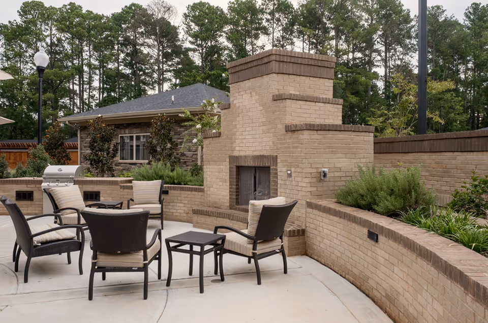 Outdoor patio area with a brick fireplace, surrounded by cushioned wicker chairs and a small table. There is a built-in grill on the left side and greenery planted along the curved brick wall. Trees and a building are visible in the background.