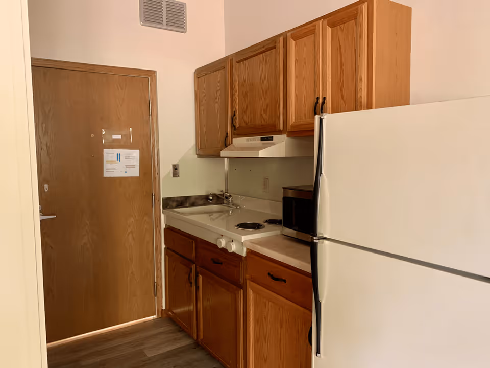 Small kitchen area with wooden cabinets, a white refrigerator, a microwave, a stove with two burners, and a sink. A wooden door is visible to the left with some papers taped to it. The floor has wood-like vinyl flooring.