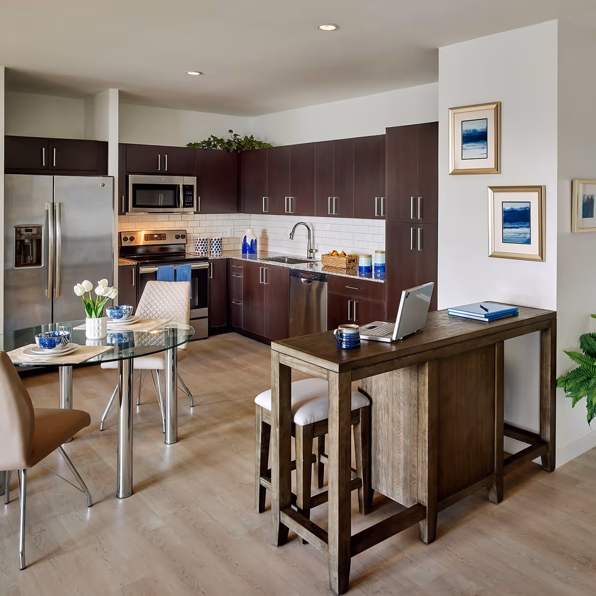 Modern kitchen with dark wood cabinets, stainless steel refrigerator, oven, microwave, and dishwasher. A glass dining table with beige chairs is set with blue and white bowls and plates. A wooden high table with two stools holds a laptop, a mug, and some books. The walls are decorated with framed blue and white artwork, and there is a green plant in the corner.