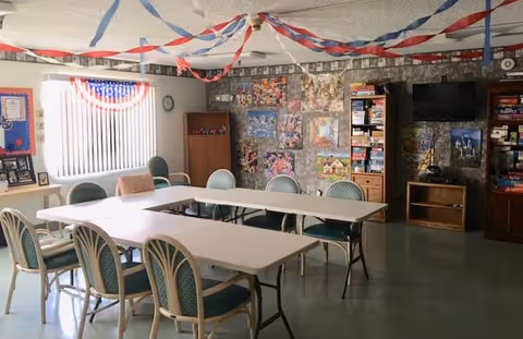 A community room decorated with red, white, and blue streamers hanging from the ceiling. There are several tables arranged in a U-shape with chairs around them. The walls feature a bulletin board, a clock, a window with vertical blinds, and a display of board games and puzzles. A television is mounted on the wall above a small wooden cabinet.