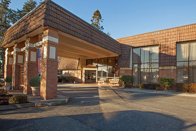 Front entrance of a brick senior living facility with a covered porte-cochère, large windows, potted shrubs, and an American flag.