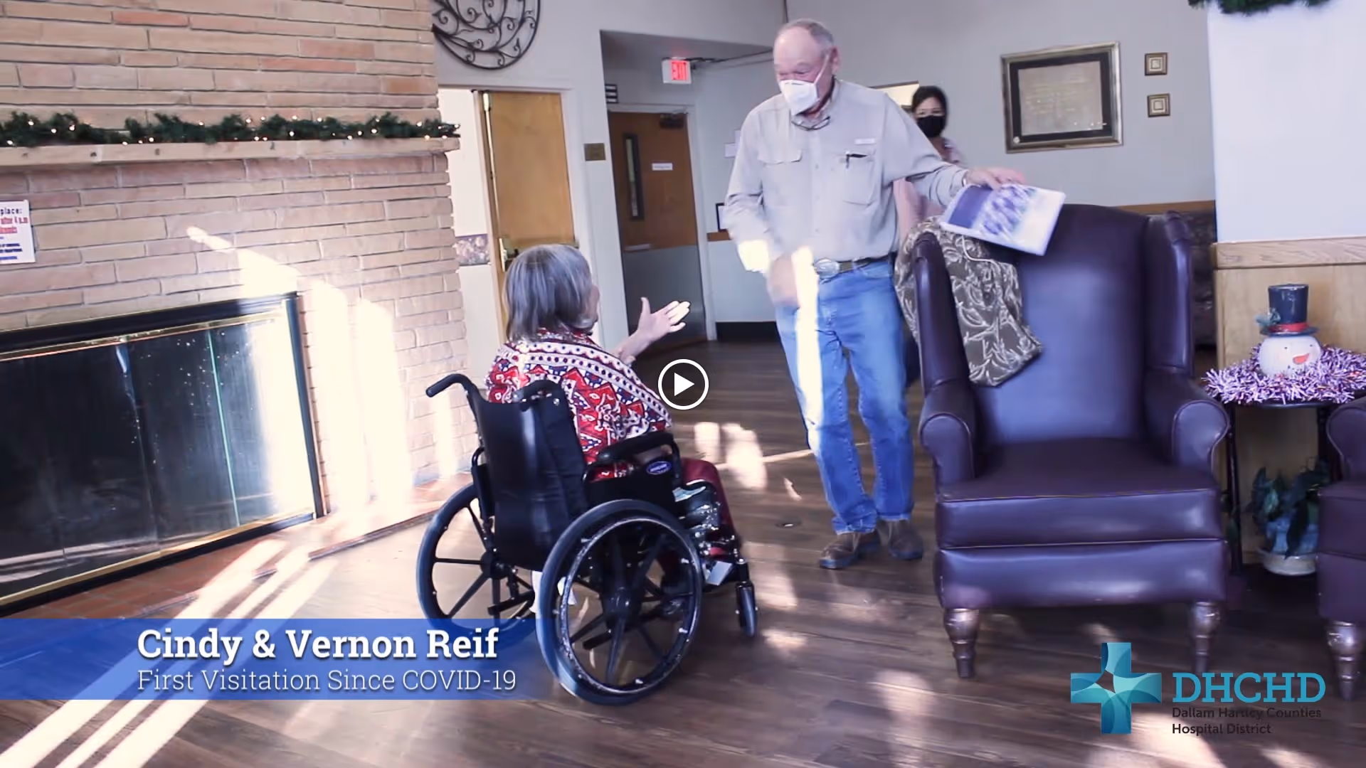 An elderly woman in a wheelchair wearing a red patterned sweater is interacting with an elderly man wearing a face mask and holding a magazine in a room with wooden floors, a brick fireplace, and leather armchairs. A woman wearing a face mask stands in the background. The room is decorated with holiday garland and a snowman decoration on a side table.