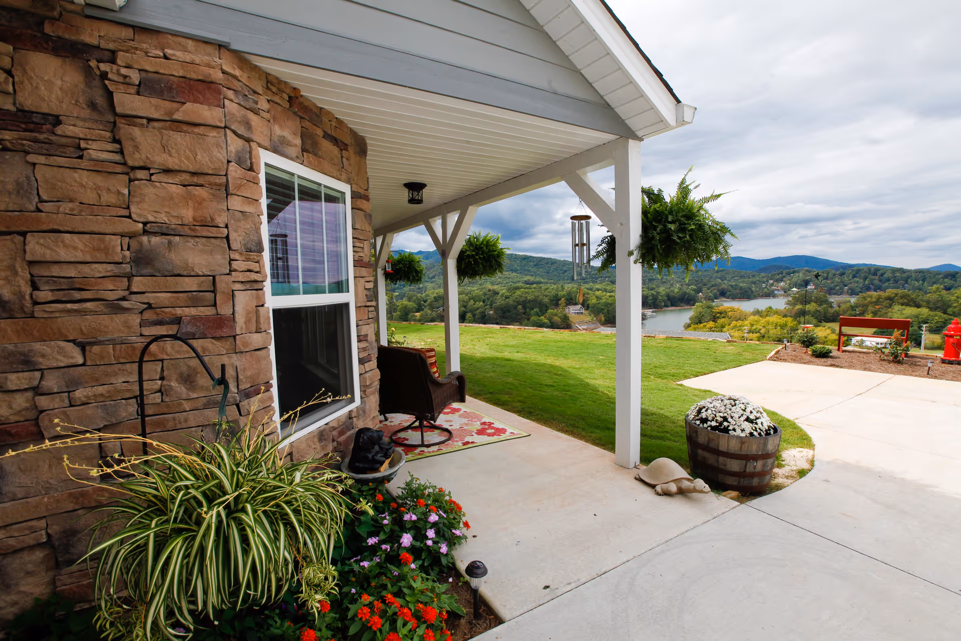 Covered patio area with stone exterior wall, a window, hanging plants, a wicker chair on a floral rug, and various potted plants. The patio overlooks a grassy lawn, a lake, and forested hills under a cloudy sky.