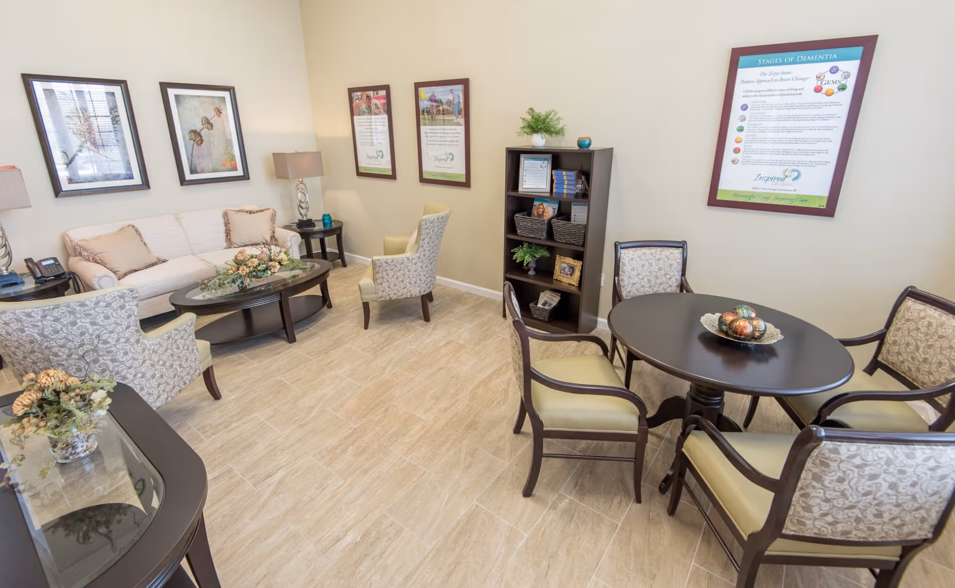A well-lit living room area in a senior living facility featuring a beige sofa with cushions, two patterned armchairs, a dark wood oval coffee table with a floral centerpiece, and two side tables with lamps. There is a round dark wood table with four upholstered chairs, a dark wood bookshelf with decorative items, and framed informational posters on the walls. The floor is tiled with light-colored tiles.