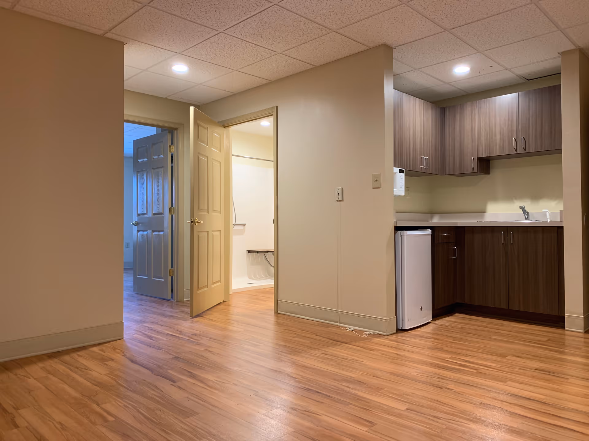 Interior view of a senior living facility showing a small kitchenette with wooden cabinets, a mini refrigerator, and a sink. To the left, there is an open door leading to a bathroom with a shower and grab bars. Another door in the background leads to another room with natural light coming through a window.