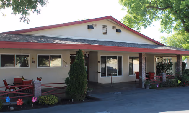 Exterior view of a single-story building with a covered porch area. The building has beige walls, a red roof trim, and several windows. There are outdoor seating arrangements with red chairs and tables on the porch. Small decorative flowers and plants are placed near the entrance, and large trees provide shade.