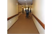 Long interior hallway with handrails and a caregiver pushing a resident in a wheelchair away from the camera.