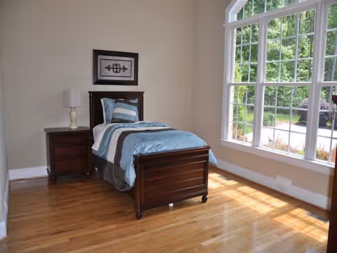 A bright bedroom with a single wooden bed dressed in blue and white bedding, a matching wooden nightstand with a lamp, a framed artwork on the beige wall, and a large window letting in natural light overlooking greenery outside.
