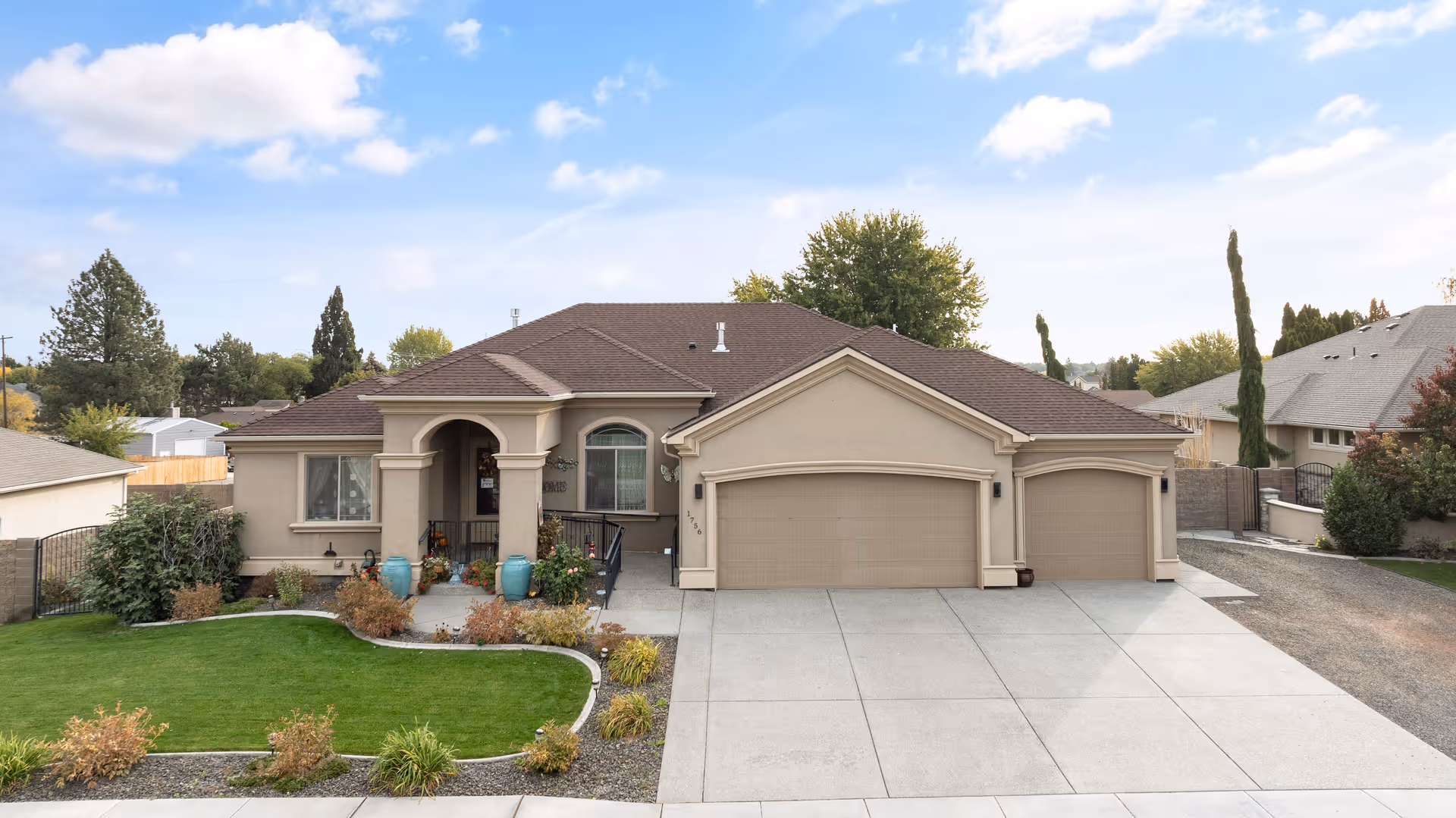 Front exterior view of a single-story beige house with a brown roof, a three-car garage, a small front porch with two blue pots, and a well-maintained lawn with shrubs and plants. The sky is partly cloudy.