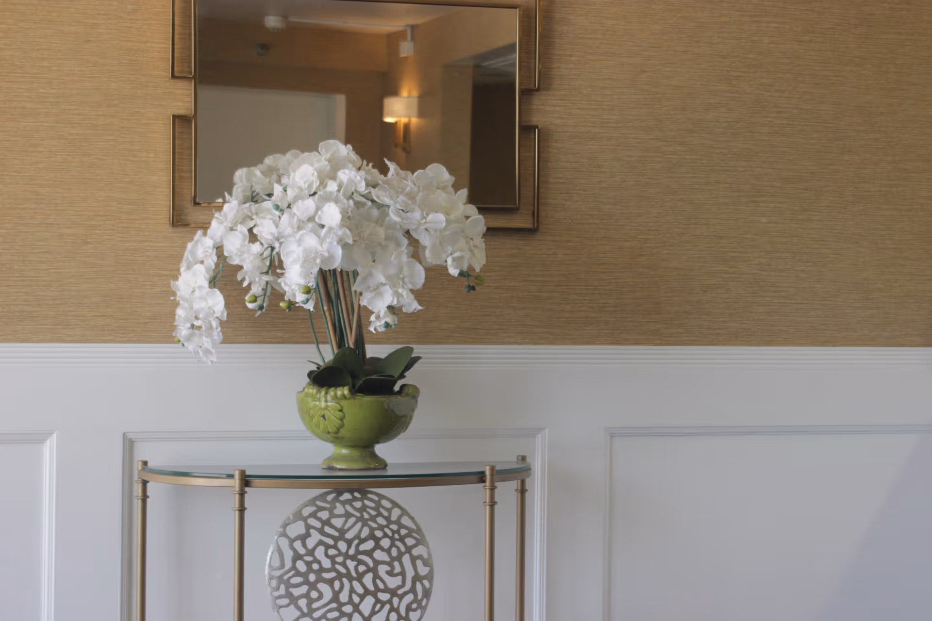 Decorative entry table with a green vase of white orchids beneath a mirror on a beige and white paneled wall.