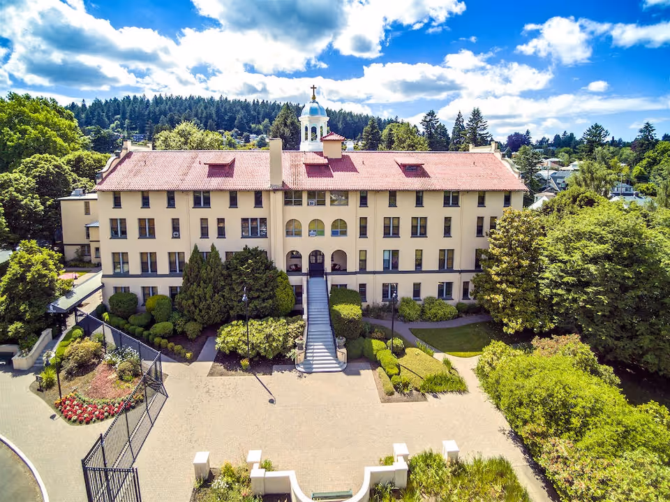 Front exterior view of a large, historic building with a red-tiled roof and a central cupola topped with a cross. The building is surrounded by well-maintained gardens and trees under a partly cloudy blue sky.