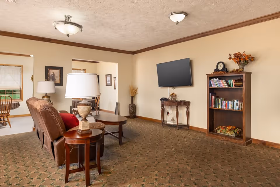 A cozy living room area with a brown recliner sofa, two wooden side tables with lamps, a wall-mounted flat screen TV, a wooden bookshelf filled with books and decorative items, and a small wooden table beneath the TV. The room has beige walls, a patterned carpet, and ceiling lights. In the background, there is a dining area with wooden chairs and a window with white curtains.