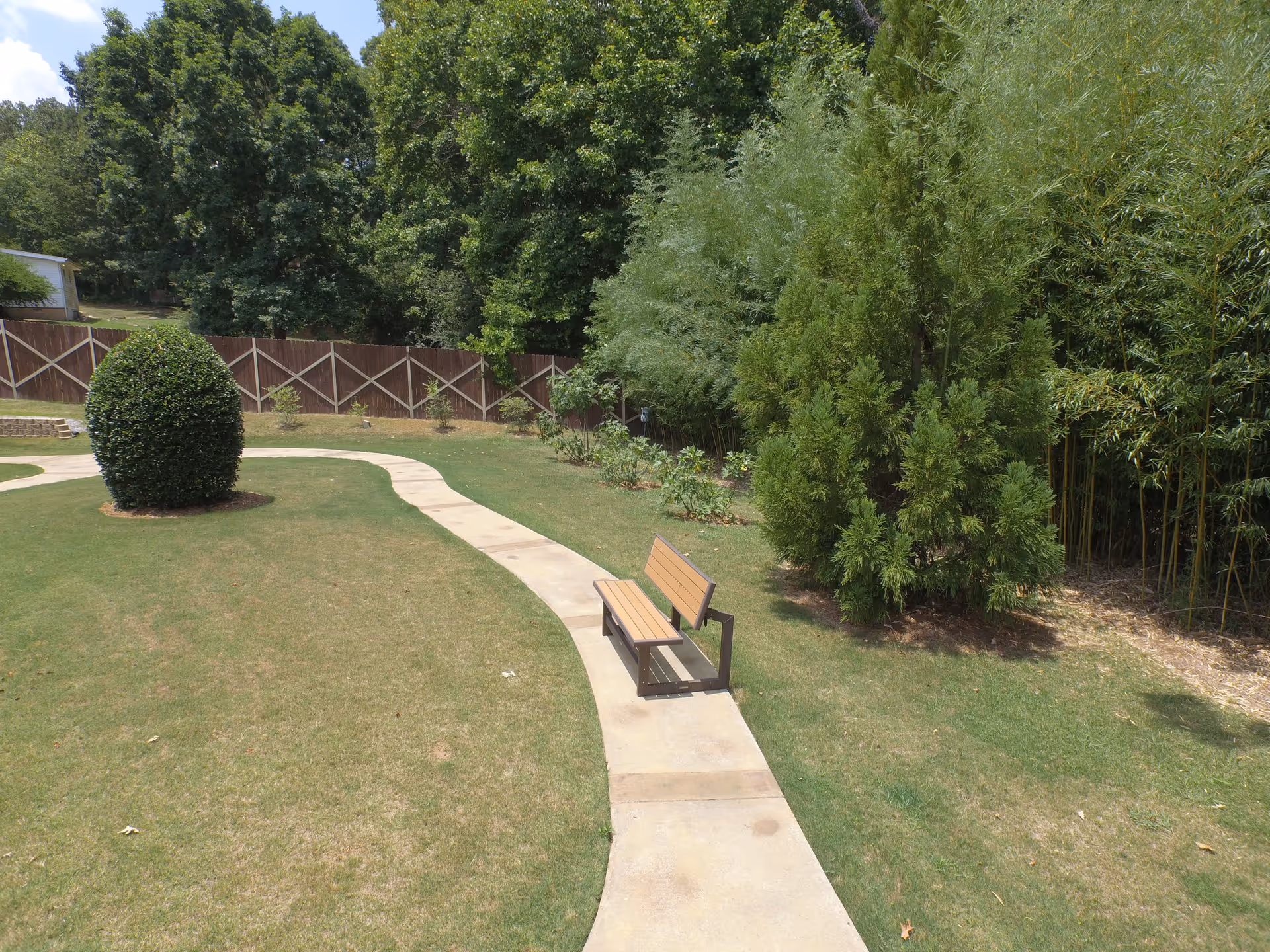 A curved concrete pathway in a grassy garden area with a wooden bench on the right side. The garden is surrounded by various green trees and bushes, with a wooden fence in the background under a partly cloudy sky.