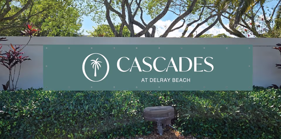 Outdoor view of a green sign mounted on a white wall with the text 'Cascades at Delray Beach' and a palm tree logo, surrounded by green bushes and trees under a blue sky.