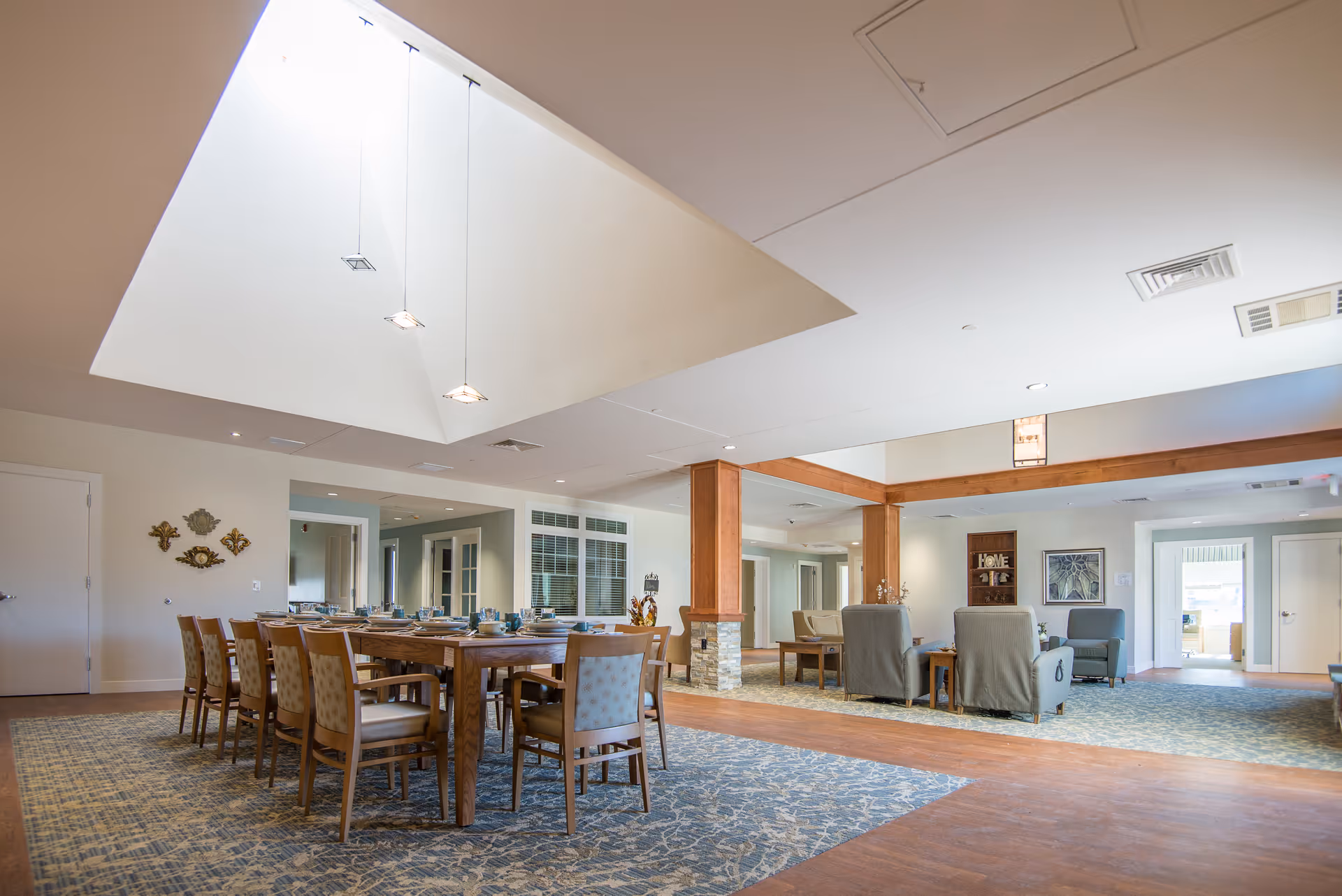 A spacious and well-lit common area in a senior living facility featuring a large wooden dining table with chairs set for a meal on the left side, and a comfortable seating area with armchairs and coffee tables on the right side. The room has a high ceiling with hanging pendant lights and a skylight, carpeted floors with a patterned design, and wooden beams dividing the space.