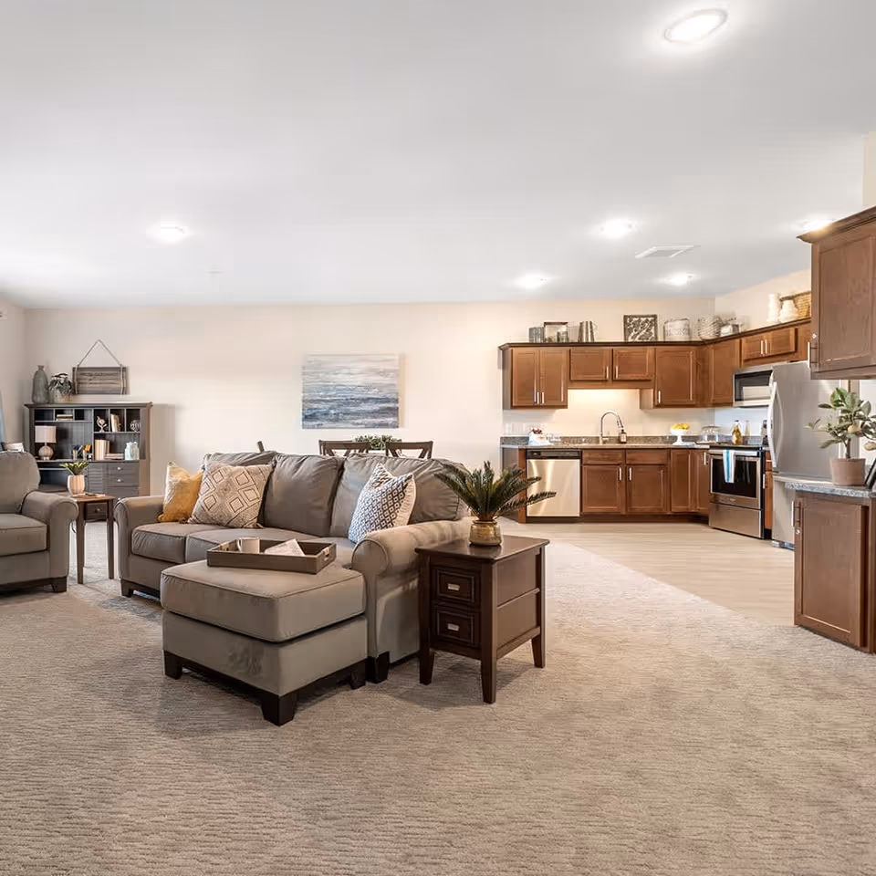 Open concept living area in a senior living facility featuring a beige sectional sofa with patterned pillows, a matching armchair, a wooden side table with a plant, a dining table with chairs, and a kitchen with wooden cabinets, stainless steel appliances, and granite countertops.