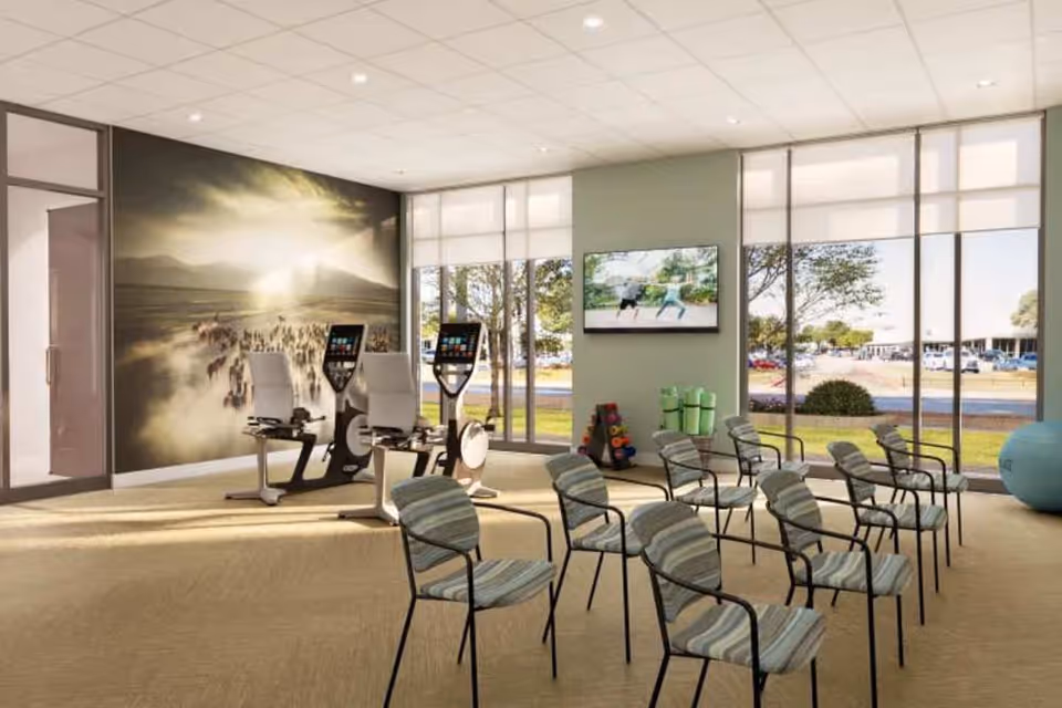 Bright activity/fitness room with rows of striped chairs, two recumbent exercise bikes, a wall mural, a TV, and large windows looking out to the parking lot.
