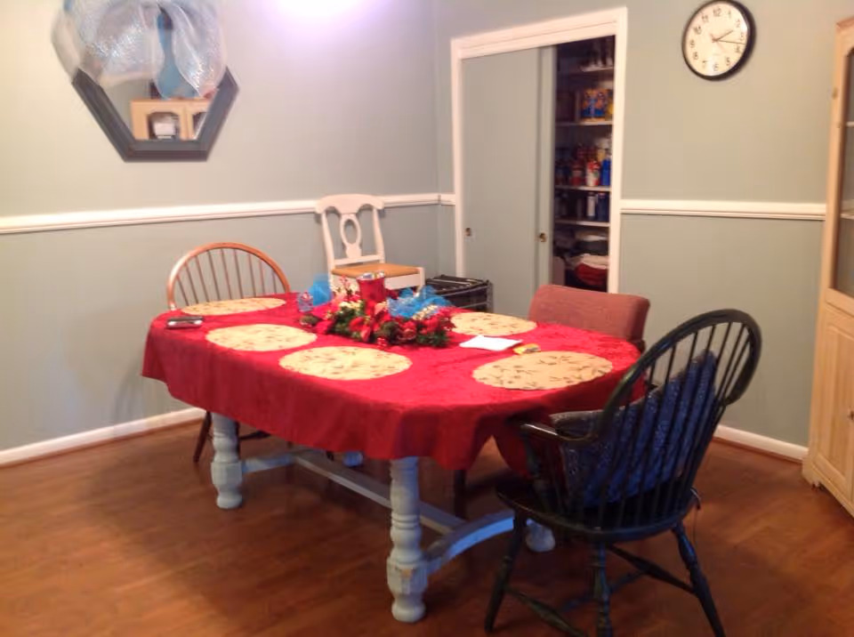 A dining room with a rectangular table covered with a red tablecloth and six round placemats. The table has a centerpiece with flowers and decorative items. There are four chairs around the table, each different in style. The room has light green walls with white trim, a clock on the wall, a mirror, and a cabinet. A pantry with sliding doors is visible in the background.