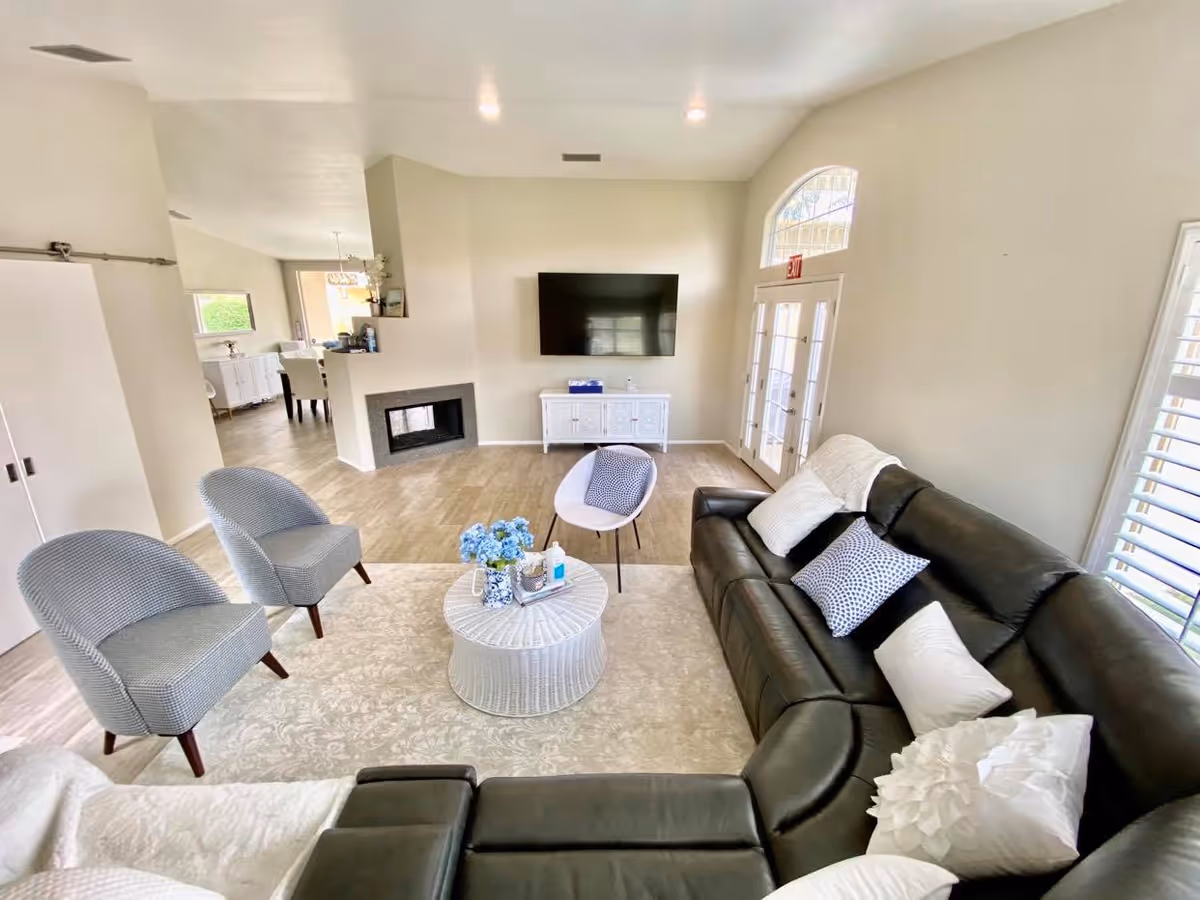 A bright and spacious living room with a black leather sectional sofa adorned with white and patterned pillows, two gray upholstered chairs, and a white round coffee table with a blue flower arrangement. A flat-screen TV is mounted on the wall above a white cabinet. The room has light-colored walls, wood flooring, a fireplace, and large windows with white shutters allowing natural light to enter. French doors with an exit sign above them are visible on the right side.