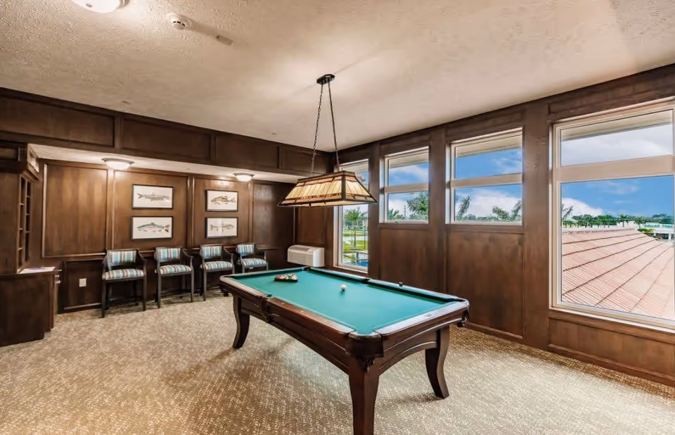 A well-lit game room with a pool table in the center, four striped cushioned chairs lined up against a wood-paneled wall, framed fish artwork, large windows showing a view of trees and a red roof outside, and a hanging light fixture above the pool table.