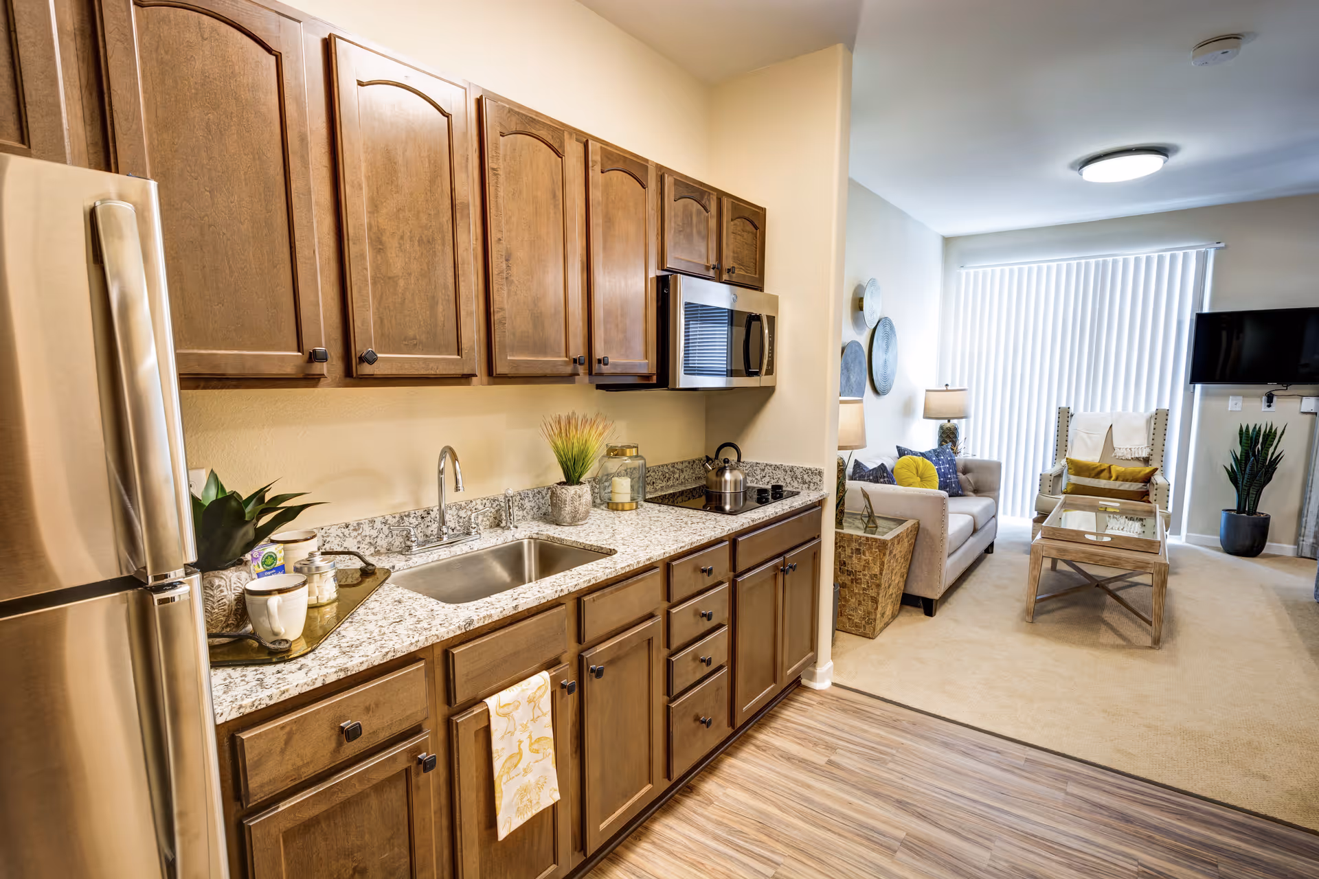 A modern kitchen with wooden cabinets, granite countertops, a stainless steel refrigerator, microwave, and sink. The kitchen opens into a living room area with a sofa, armchair, coffee table, floor lamp, and a wall-mounted TV. The living room has a large window with vertical blinds and a potted plant in the corner.