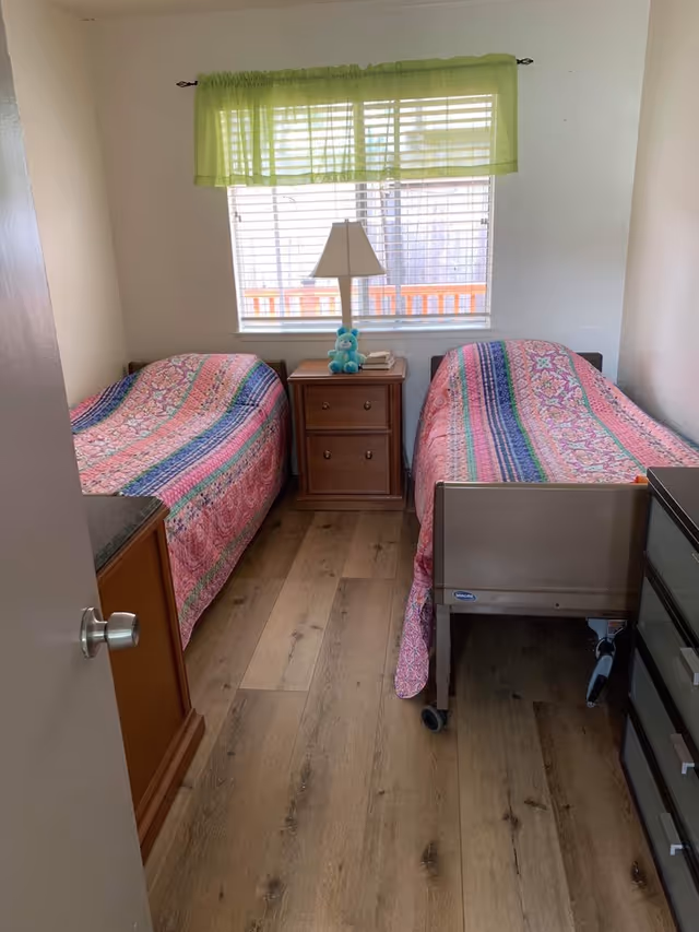 A small bedroom with two single beds covered in colorful striped quilts. Between the beds is a wooden nightstand with a lamp, a blue stuffed animal, and some books. A window with white blinds and a green valance is above the nightstand. The floor is wooden, and there is a door partially open on the left side.